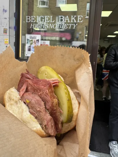 A salt-beef beigel with a pickle held in front of the Beigel Bake shop in London