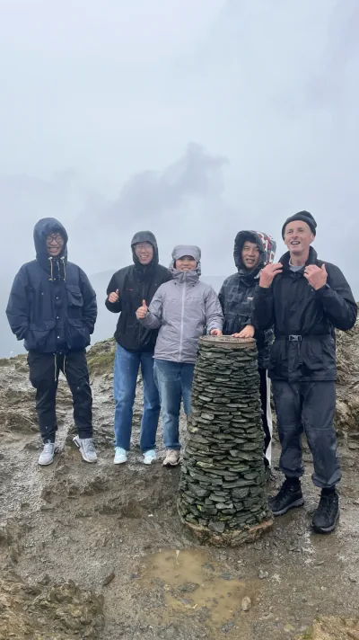 Five hikers gathered around a stone summit marker on a misty mountain near Keswick