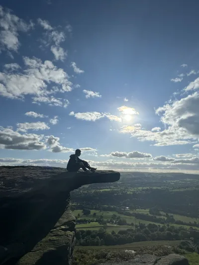A seated figure silhouetted on a rocky ledge above the Peak District under a bright, cloud-filled sky